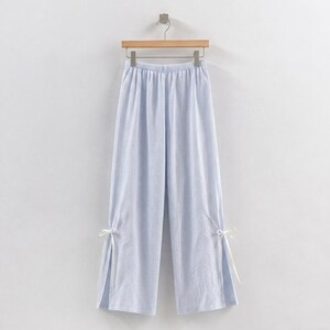 Puede incluir: Pantalones de pernera ancha a rayas azules y blancas, colgados de una percha de madera. Los pantalones tienen cintura con cord&oacute;n y aberturas en los tobillos, atadas con cintas blancas. La tela parece ligera y veraniega.