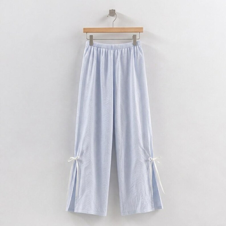 Puede incluir: Pantalones de pernera ancha a rayas azules y blancas, colgados de una percha de madera. Los pantalones tienen cintura con cord&oacute;n y aberturas en los tobillos, atadas con cintas blancas. La tela parece ligera y veraniega.
