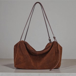 May include: A brown suede shoulder bag with long, thin brown straps. The bag has a soft, slouchy shape and a simple design. The straps are attached with silver-colored hardware. The bag is sitting on a light-colored surface.