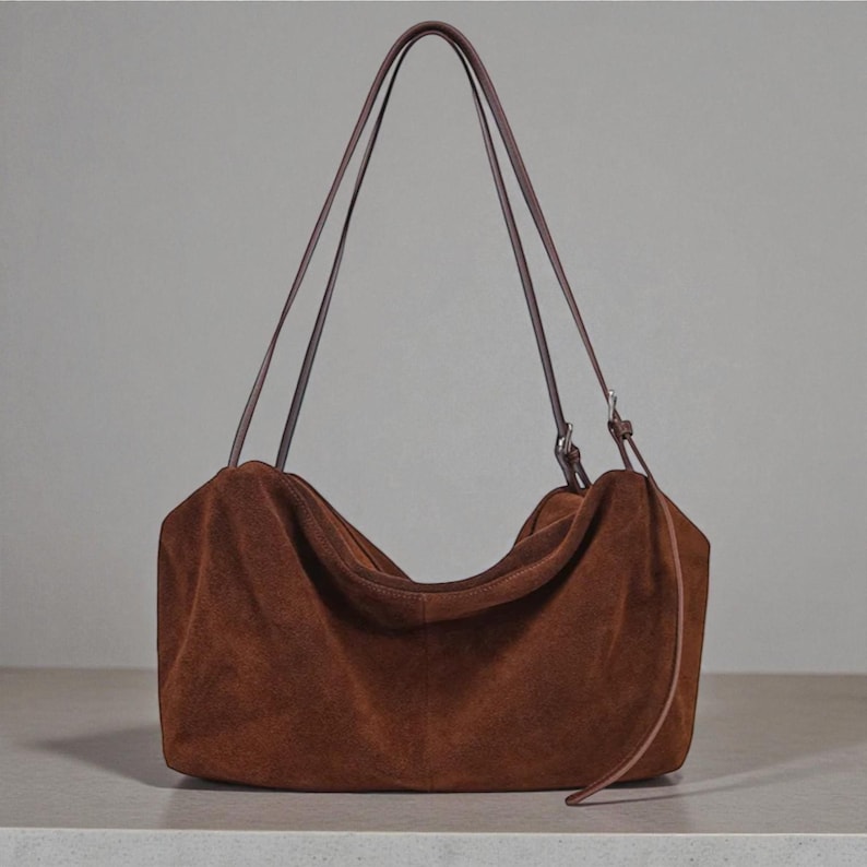 May include: A brown suede shoulder bag with long, thin brown straps. The bag has a soft, slouchy shape and a simple design. The straps are attached with silver-colored hardware. The bag is sitting on a light-colored surface.