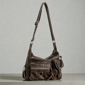 May include: A brown leather shoulder bag with multiple zippered pockets and a long adjustable strap. The bag features a D-ring detail and a small tassel. The bag is made of a soft, worn leather.