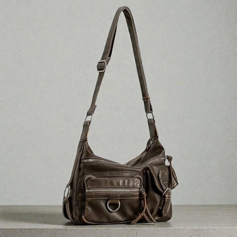 May include: A brown leather shoulder bag with multiple zippered pockets and a long adjustable strap. The bag features a D-ring detail and a small tassel. The bag is made of a soft, worn leather.