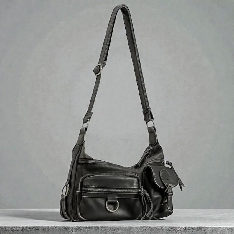 May include: Black leather shoulder bag with multiple exterior pockets and a long adjustable strap. The bag features silver-tone hardware, including D-rings and buckles. The bag has a zippered main compartment and several smaller pockets.