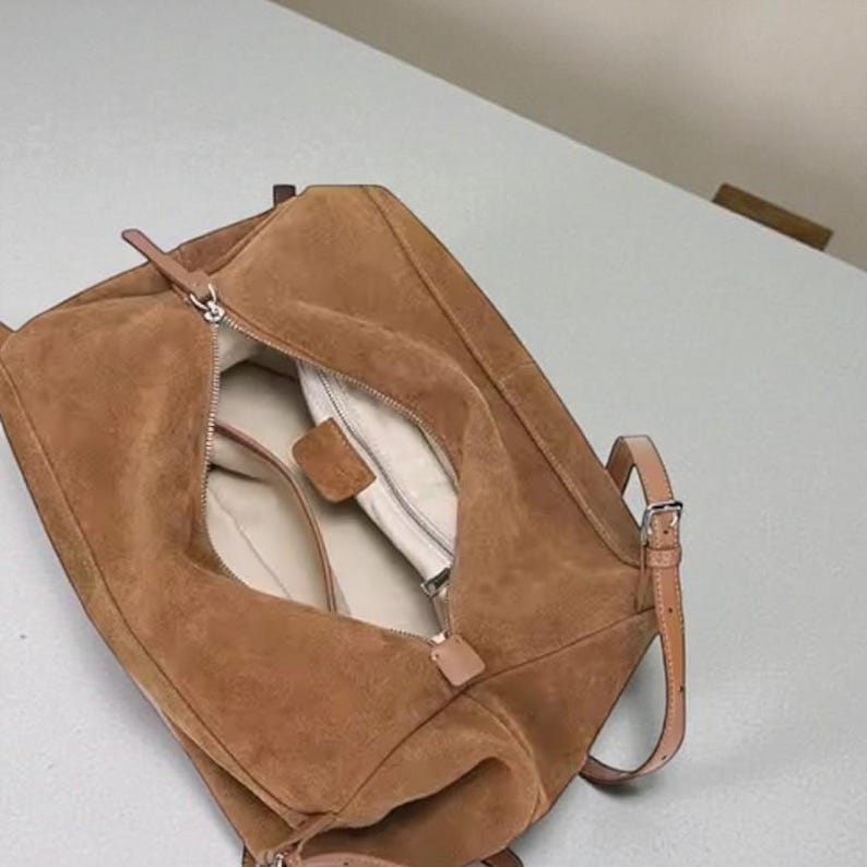 May include: A brown suede shoulder bag with a zipper closure. The bag has a rectangular shape with a top handle and a long adjustable strap. The interior is lined with a light-colored fabric.