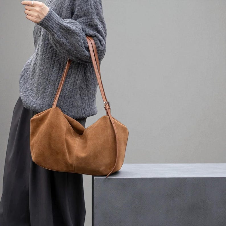 May include: A brown suede shoulder bag with tan leather straps and a buckle closure. The bag has a slouchy, unstructured design and is carried over the shoulder. The bag is a warm, earthy tone and is set against a neutral background.