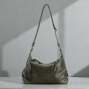 May include: Olive green shoulder bag with a long, adjustable strap. The bag has a curved shape, a zipper closure, and a small zipper pocket. The bag is made of a soft, textured material and has a minimalist design.