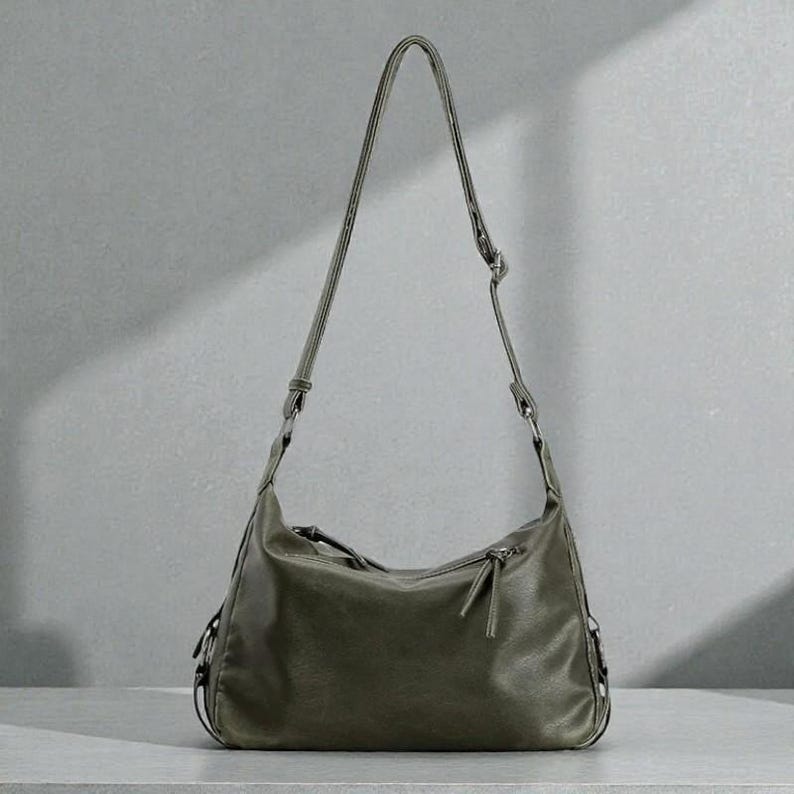 May include: Olive green shoulder bag with a long, adjustable strap. The bag has a curved shape, a zipper closure, and a small zipper pocket. The bag is made of a soft, textured material and has a minimalist design.