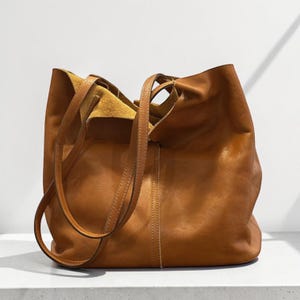 May include: A tan leather tote bag with a soft, unstructured silhouette, featuring two long shoulder straps and a vertical stitched seam down the centre. The interior lining is visible at the top opening, showing a lighter, natural suede texture.