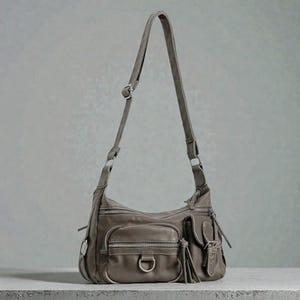 May include: A gray leather shoulder bag with multiple zippered pockets and a long adjustable strap. The bag features a D-ring detail and decorative tassels, offering a stylish and functional design. The bag is sitting on a gray surface.