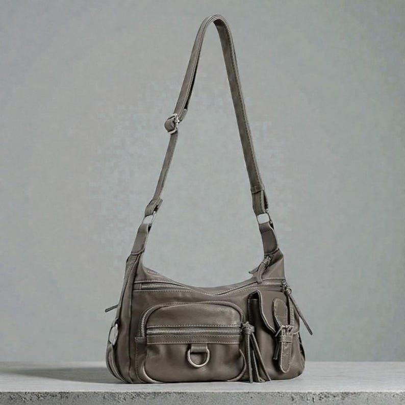May include: A gray leather shoulder bag with multiple zippered pockets and a long adjustable strap. The bag features a D-ring detail and decorative tassels, offering a stylish and functional design. The bag is sitting on a gray surface.