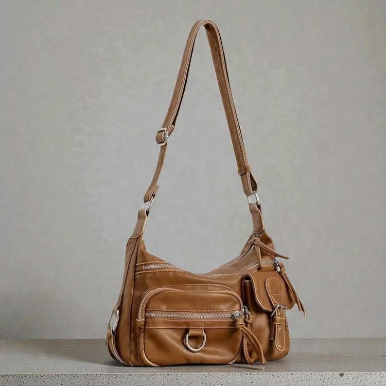 May include: A brown leather shoulder bag with multiple exterior pockets and silver-toned hardware. The bag features a long, adjustable strap and a zip closure on each pocket. The bag has a tassel detail on one of the pockets.
