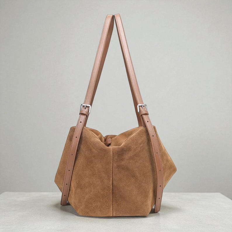May include: A brown suede handbag with tan leather straps and silver-tone buckles. The bag has a soft, slouchy design and a rectangular base. The straps are adjustable and extend upwards, creating a unique silhouette.