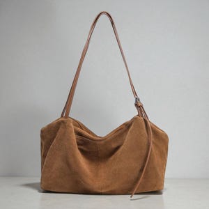 May include: A brown suede shoulder bag with a long, thin strap. The bag has a rectangular shape with a gathered top and a drawstring detail. The strap is a lighter brown color than the bag.