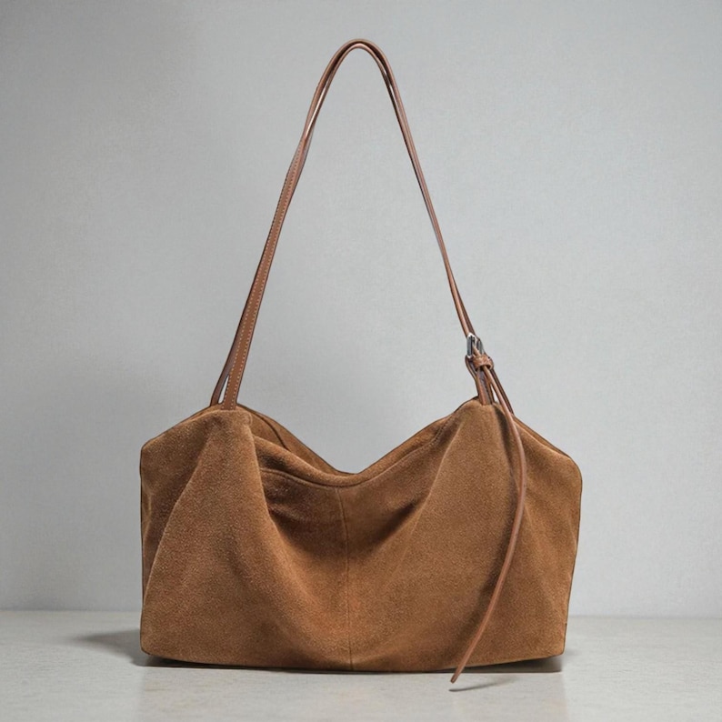May include: A brown suede shoulder bag with a long, thin strap. The bag has a rectangular shape with a gathered top and a drawstring detail. The strap is a lighter brown color than the bag.