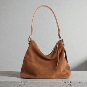 May include: A brown suede shoulder bag with a curved shape and a long, light brown leather strap. The bag has a silver zipper and a matching leather pull. The bag is sitting on a gray surface.