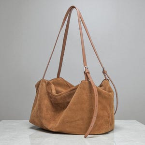 May include: A large, brown suede tote bag with long, thin tan leather straps. The bag has a slouchy, unstructured design and a silver-colored buckle on the strap. The bag is sitting on a white surface.