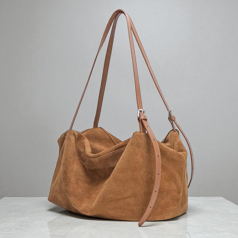 May include: A large, brown suede tote bag with long, thin tan leather straps. The bag has a slouchy, unstructured design and a silver-colored buckle on the strap. The bag is sitting on a white surface.