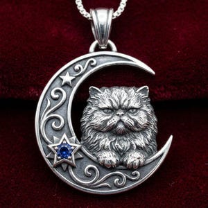 May include: A sterling silver pendant featuring a detailed cat face centered within a crescent moon. The moon is decorated with swirling filigree patterns, a small plain star, and a larger star set with a single round, faceted blue gemstone.