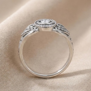 May include: A silver ring with an oval setting, a central clear gemstone, and surrounding smaller stones. The band features detailed, swirling patterns and decorative elements on the shoulders, giving it a vintage or antique appearance.