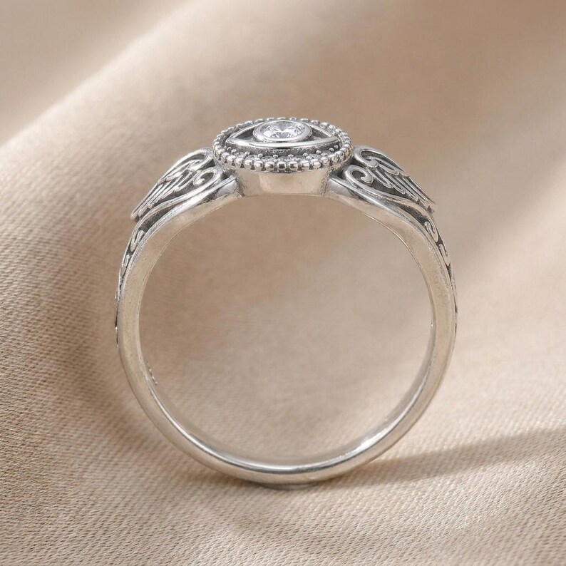 May include: A silver ring with an oval setting, a central clear gemstone, and surrounding smaller stones. The band features detailed, swirling patterns and decorative elements on the shoulders, giving it a vintage or antique appearance.