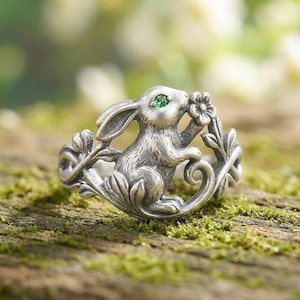 May include: A silver ring featuring a sculpted rabbit figure integrated into a leafy vine design. The rabbit has a small, bright green gemstone set as its eye. The metal has an oxidized finish that highlights the detailed texture of the fur and leaves.