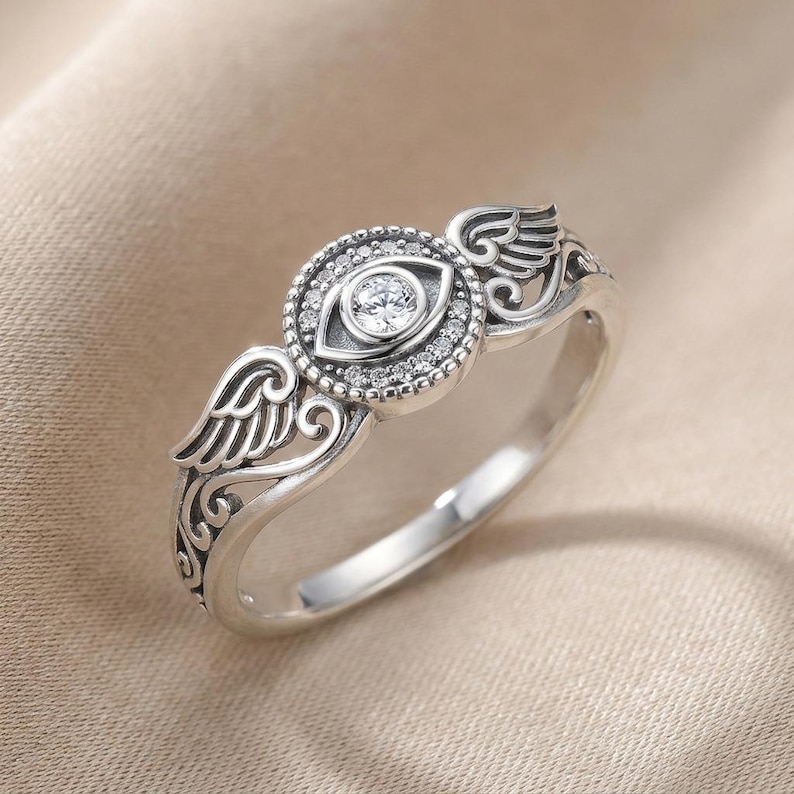 May include: A silver ring with an eye motif, a central clear stone, and smaller surrounding stones. The ring features wing-shaped embellishments on either side of the eye, with detailed patterns.