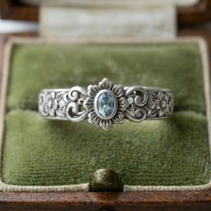 May include: A silver ring with an oval, light blue gemstone set in a floral design. The band features intricate floral and scrollwork patterns. The ring is displayed in a green velvet jewelry box.