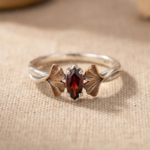 May include: A silver ring featuring a central marquise-cut dark red garnet gemstone. The band is accented on both sides of the stone with detailed, fan-shaped ginkgo leaf motifs in a contrasting bronze-toned metal finish with textured veins.