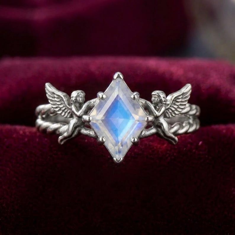 May include: A silver ring featuring a central kite-shaped moonstone with a blue iridescent glow. The band is designed with a twisted rope texture and two winged cherub figures positioned on either side of the gemstone, facing inward.
