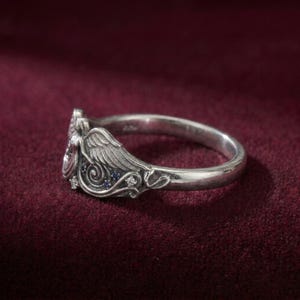 May include: A silver ring with an angel wing design. The ring features small blue and clear gemstones. The ring is set against a dark red background.