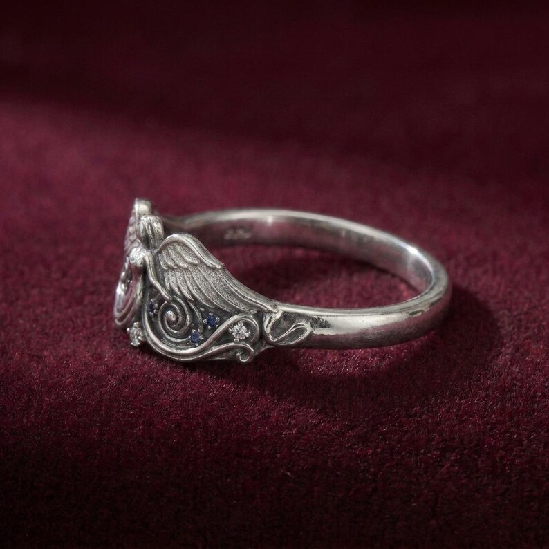 May include: A silver ring with an angel wing design. The ring features small blue and clear gemstones. The ring is set against a dark red background.