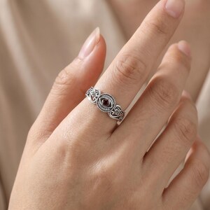 May include: A silver ring with an eye motif, a central red gemstone, and surrounding clear stones. The ring has an intricate design with wing-shaped details on either side of the eye.