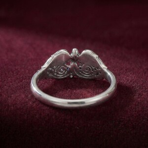 May include: A silver ring with an ornate design featuring an eagle or bird motif. The ring has a detailed, openwork pattern with small blue gemstones. The ring is set against a dark red background.