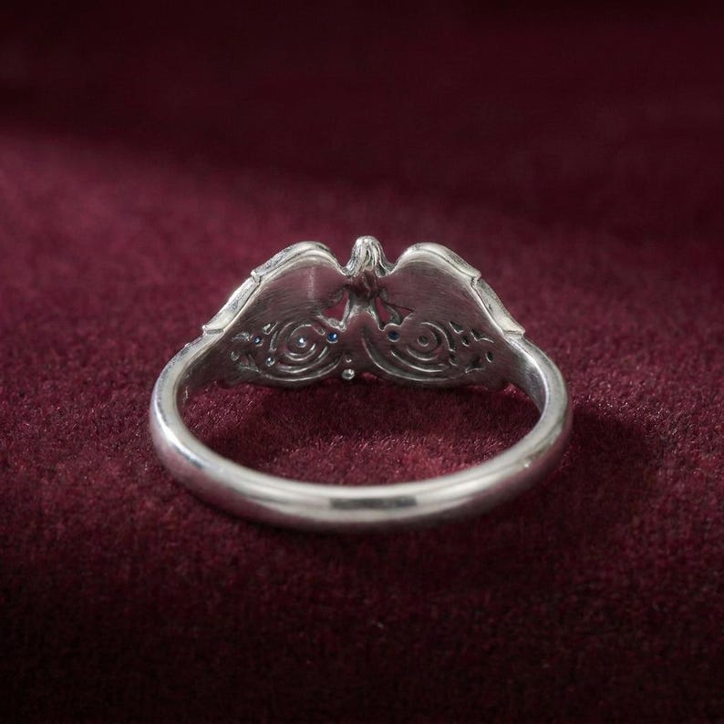 May include: A silver ring with an ornate design featuring an eagle or bird motif. The ring has a detailed, openwork pattern with small blue gemstones. The ring is set against a dark red background.