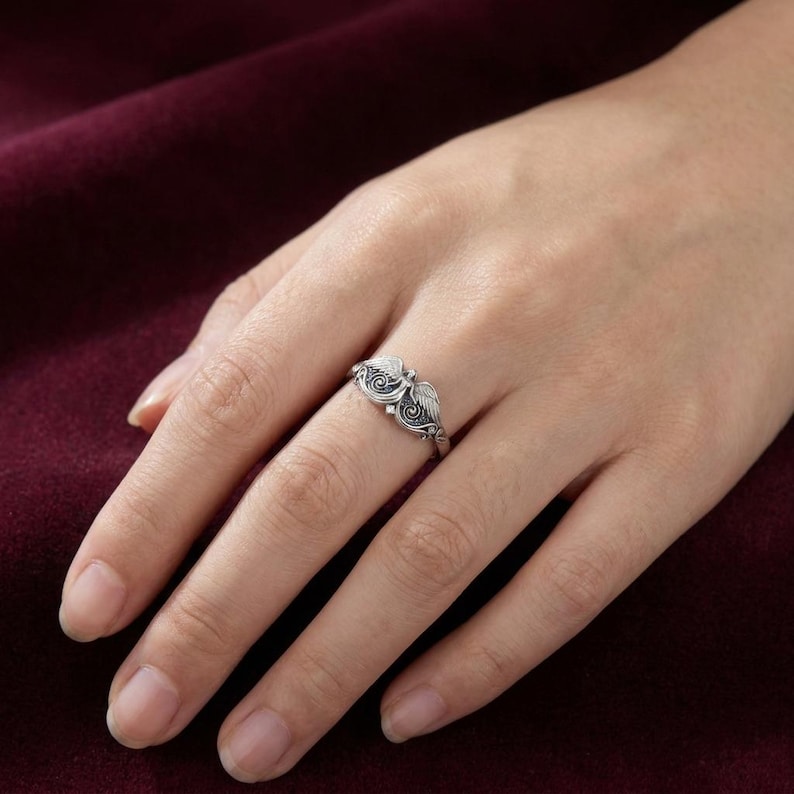 May include: A silver ring with an ornate design featuring two stylized wings. The ring is worn on a finger, showcasing the intricate details of the metalwork. The ring's design includes swirling patterns and a central element.
