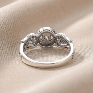 May include: A silver ring with a central, round, clear gemstone surrounded by smaller stones. The ring features wing-like designs on either side of the central stone. The band is smooth and polished, with decorative details near the wings.