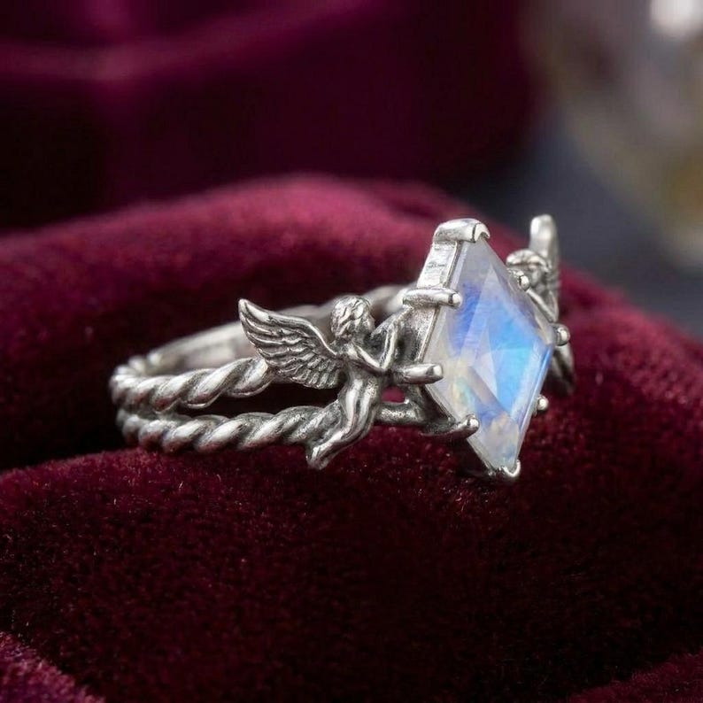 May include: A silver ring featuring a central kite-shaped moonstone gemstone. The band has a twisted rope texture and is flanked by two detailed angel figures with outstretched wings that frame the iridescent blue and white stone setting.