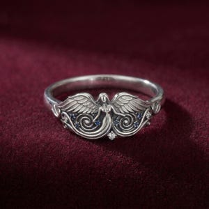 May include: A silver ring featuring an angel design. The ring has an angel figure with outstretched wings, accented with small blue and clear gemstones. The ring is set against a deep red background.