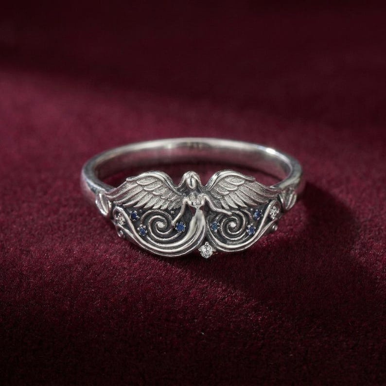 May include: A silver ring featuring an angel design. The ring has an angel figure with outstretched wings, accented with small blue and clear gemstones. The ring is set against a deep red background.