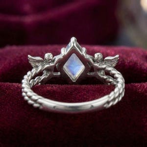 May include: A silver ring featuring a central kite-shaped iridescent moonstone. The band is designed with a twisted rope texture and two small winged cherub figures positioned on either side of the central gemstone setting.