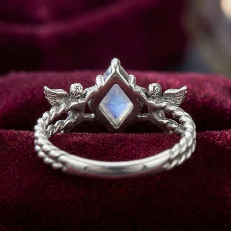 May include: A silver ring featuring a central kite-shaped iridescent moonstone. The band is designed with a twisted rope texture and two small winged cherub figures positioned on either side of the central gemstone setting.