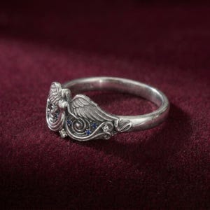 May include: A silver ring featuring an angel design with detailed wings and a flowing gown. The ring is adorned with small blue and clear gemstones, adding sparkle to the angelic figure. The ring is set against a deep red background.