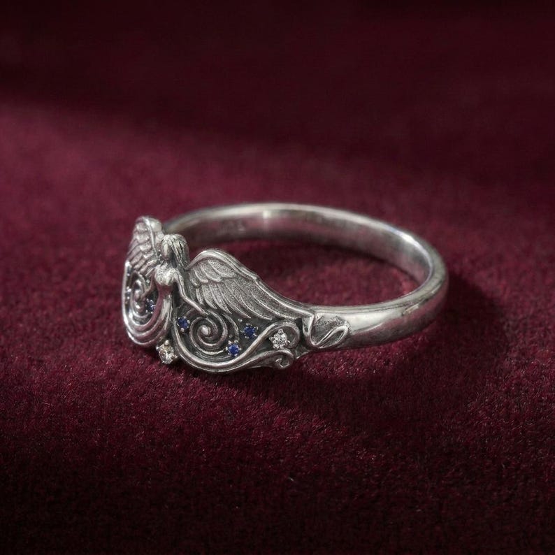 May include: A silver ring featuring an angel design with detailed wings and a flowing gown. The ring is adorned with small blue and clear gemstones, adding sparkle to the angelic figure. The ring is set against a deep red background.