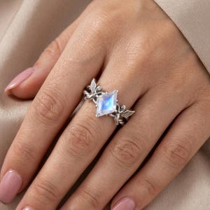 May include: A silver ring featuring a central kite-shaped iridescent moonstone. The band is crafted with detailed metalwork depicting two small winged figures on either side of the stone, creating a symmetrical design with a polished finish.