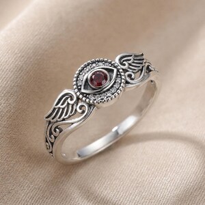 May include: A silver ring with an eye motif, a central red stone, and surrounding clear stones. The ring features wing details on either side of the eye and detailed scrollwork.