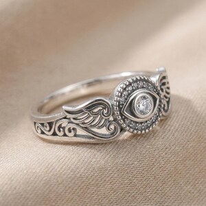 May include: A silver ring with an eye motif, set with a central clear stone and smaller surrounding stones. The ring features wing and scroll detailing on the band. The ring is presented on a neutral-coloured surface, showcasing its intricate design.