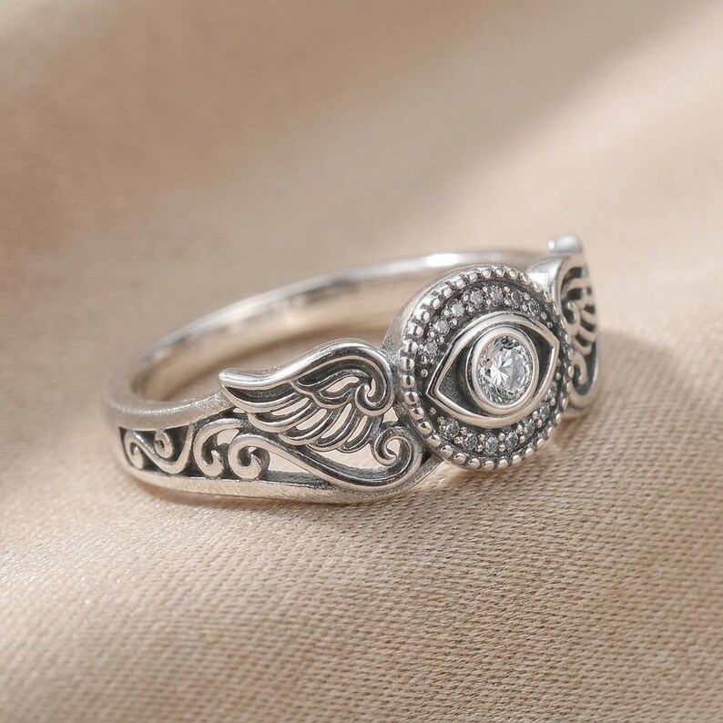 May include: A silver ring with an eye motif, set with a central clear stone and smaller surrounding stones. The ring features wing and scroll detailing on the band. The ring is presented on a neutral-coloured surface, showcasing its intricate design.