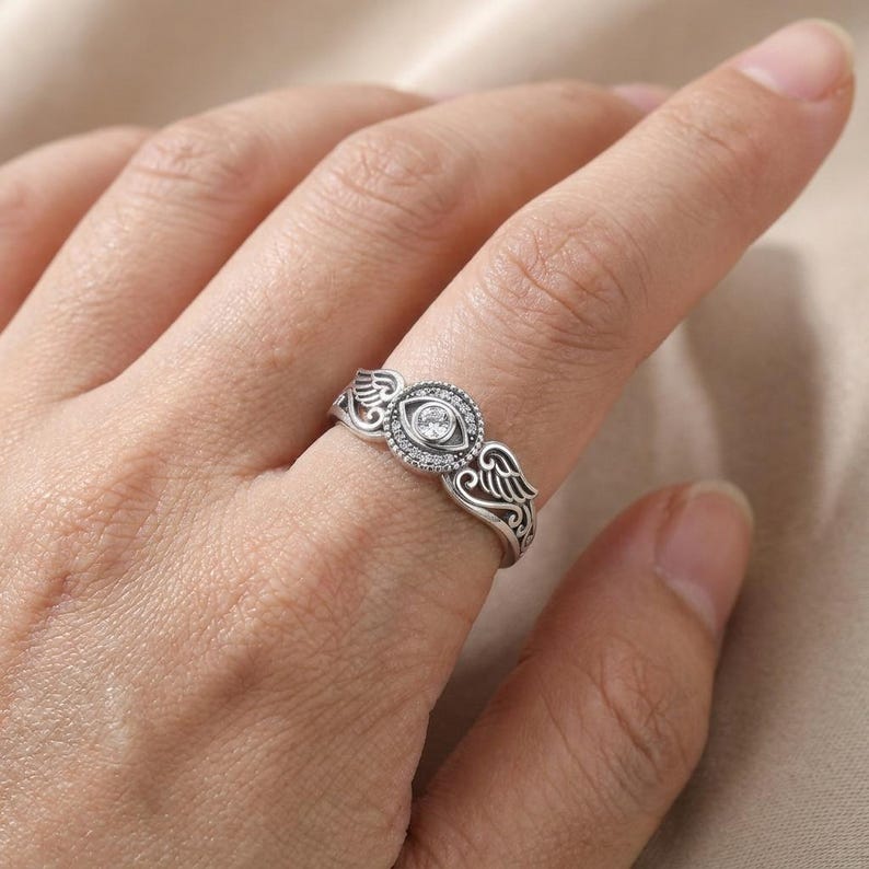May include: A silver ring with an eye motif, a central clear stone, and surrounding smaller stones. The ring has wing-shaped embellishments on either side of the eye. A distinctive piece of jewellery with symbolic design.