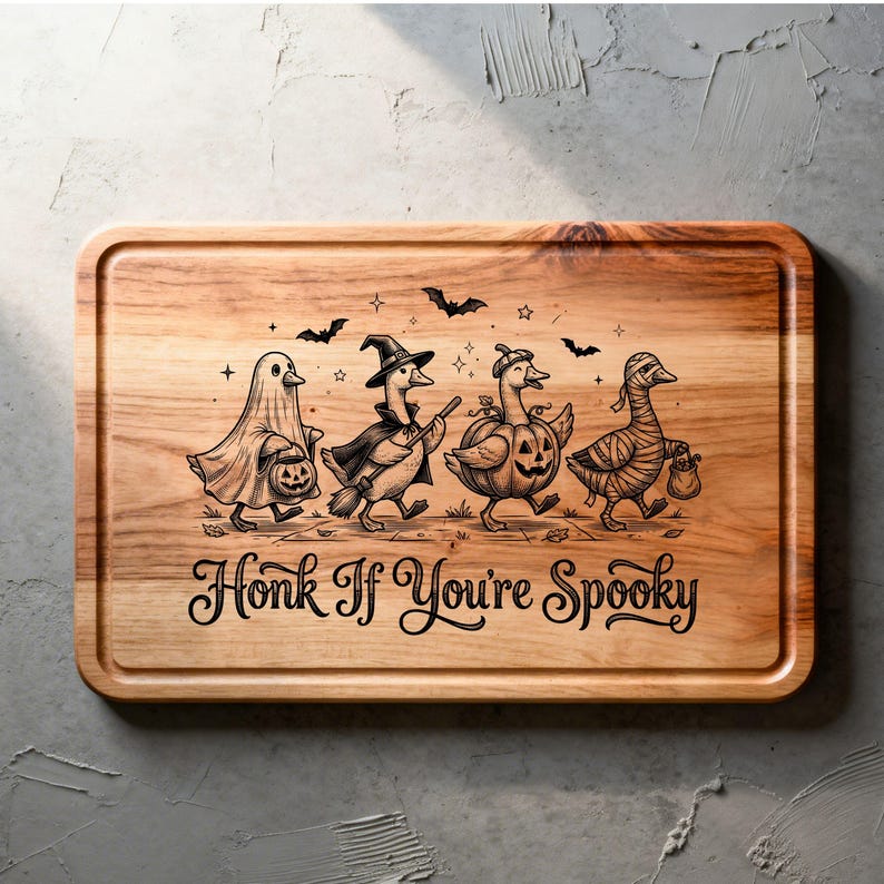 May include: A rectangular wooden cutting board featuring a laser-engraved illustration of four geese in Halloween costumes: a ghost, a witch, a pumpkin, and a mummy. The text "Honk If You're Spooky" is engraved below the geese with small bats and stars.