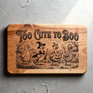 May include: A rectangular wooden cutting board featuring a laser-engraved illustration of four ducks in Halloween costumes. The text "TOO CUTE TO BOO" is centered at the top. The ducks are dressed as a ghost, a witch, a pumpkin, and a mummy.